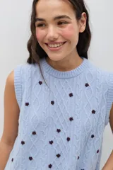 Chaleco de punto color celeste, con diseño de trenzas y pequeñas flores bordadas a mano en color marrón oscuro. Presenta cuello redondo y terminaciones acanaladas.