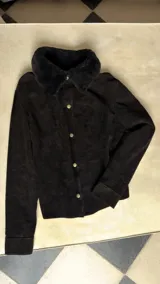 Camisa de cuero negro con cuello de corderito natural, cierre frontal con broches metálicos y puños ajustables.