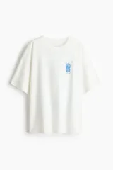 Remera oversized de algodón blanco, con cuello redondo acanalado, mangas cortas con hombros caídos y pequeño estampado azul en el pecho de un jarrón con flores y el número 1096.