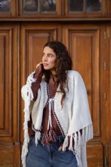 Poncho tejido de color blanco, con flecos en los bordes y detalles de bordado en crochet a lo largo de la abertura frontal. Cuenta con un cierre de cordones con borlas en el cuello.