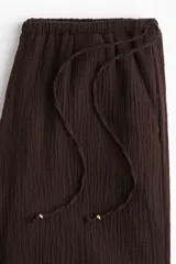Pantalón marrón de corte holgado con piernas amplias y bolsillos al bies. Modelo de tiro bajo con elástico revestido y cordones para ajustar enlazados a los lados, decorados con cuentas en los extremos.