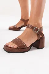 Sandalia de taco ancho y plataforma, confeccionada en cuero tostado con textura trenzada. Cuenta con una tira delantera decorada con tachas metálicas plateadas, tira en el talón con hebilla ajustable y suela con base de goma.