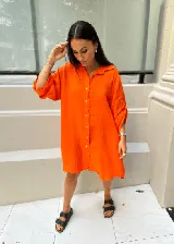 Vestido camisero corto color naranja, de lino, con cuello clásico, cierre frontal con botones y mangas 3/4 con botones para recoger. Diseño amplio y cómodo, con corte irregular más largo en la parte trasera.