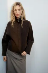 Chaqueta de punto marrón de textura bouclé, con cuello subido, manga larga ancha y cierre frontal oculto con cremallera.