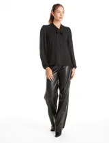 Blusa de seda fría color negro, con escote en V y lazo decorativo al cuello. Presenta mangas largas abullonadas con puños elásticos y detalles de strass aplicados en cuello, lazo y mangas.