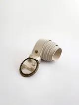 Cinto de cuero color crudo con forro de cuero y sistema de broche suizo intercambiable. Presenta una hebilla circular de guampa con borde metálico labrado en tono bronce.