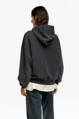 Sudadera con capucha de algodón color negro desgastado, corte globo y manga larga. Presenta un estampado frontal que muestra una escena urbana con edificios, flores amarillas y una bota de lluvia verde sobre un muro de ladrillos.