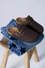 Zueco de gamuza color marrón chocolate, con detalle de tachas doradas y hebilla metálica ajustable en el empeine. Presenta una base de corcho y suela de goma antideslizante.