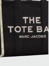 Bolso tipo tote de gran tamaño en color beige, confeccionado en lona resistente. Presenta el logo estampado en negro en el frente y detalles de cintas laterales con inscripciones. Cuenta con doble asa superior para llevar al hombro.