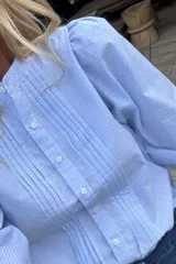 Camisa de manga larga con estampado de cuadros vichy en tonos celestes y blancos. Presenta un diseño con pliegues verticales en el frente, cuello redondo y cierre frontal con botones.