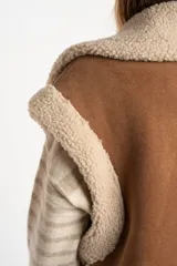 Chaleco estilo gamulán de corte oversize, confeccionado en material tipo gamuza color camel con interior y bordes de corderito sintético color crema. Presenta cierre frontal completo, bolsillos laterales y detalles de tiras decorativas en los hombros.
