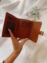 Billetera de cuero sintético con textura granulada, cierre de solapa con broche a presión y tira decorativa central.