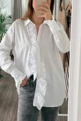 Camisa blanca de algodón con cuello clásico, cierre frontal con botones, mangas largas con puños abotonados y volados en el frente y los puños.