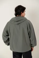 Canguro tipo anorak de color verde militar, con capucha ajustable y cierre frontal parcial. Presenta un panel central en contraste de textura gamuzada y un bolsillo frontal tipo canguro con solapa.