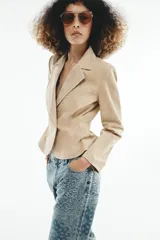 Blazer corto de cuero en color tostado, con cuello solapa, hombreras y manga larga. Presenta un diseño evasé en el bajo y un cierre frontal cruzado con dos botones.