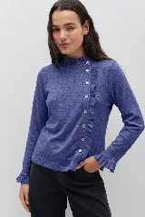 Remera de manga larga en color azul melange, con cuello alto y cierre frontal asimétrico con botones. Presenta detalles de volados a lo largo de la abotonadura, en el cuello y en los puños.