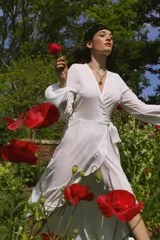 Vestido tipo batón de crepe color blanco, con escote en V, mangas largas abullonadas con puños elásticos y diseño envolvente con lazo de ajuste en la cintura. Presenta un largo irregular con volado en el ruedo.