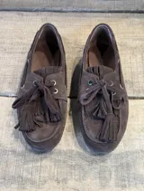 Mocasín tipo náutico de gamuza color marrón oscuro, con cordones frontales y detalle de flecos decorativos en el empeine.