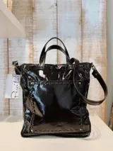 Bolso tipo tote de cuero charol negro con doble asa corta y correa larga desmontable. Presenta detalles de costuras a la vista y apliques metálicos.