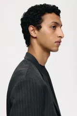 Blazer cruzado de tejido gris oscuro con finas rayas verticales blancas (pinstripe). Presenta solapas de pico, cierre de doble botonadura con botones oscuros, bolsillo superior insertado y dos bolsillos delanteros ribeteados con solapa. Tiene botones decorativos en los puños y abertura trasera.