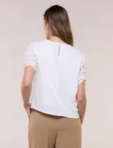Blusa blanca de corte recto con cuello redondo y mangas cortas que presentan detalles de bordado calado y terminación festoneada.
