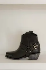 Botinetas de cuero negro con diseño estilo western, punta cuadrada y taco bajo con acabado desgastado. Presentan una tira decorativa con estribo metálico desmontable alrededor del tobillo.