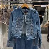 Chaqueta corta de denim estilo militar, color azul con efecto deslavado, cuello mao, hombreras y cierre frontal con múltiples filas de presillas y botones metálicos. Cuenta con mangas largas adornadas con botones en los puños.