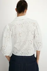 Camisa de cuello solapa y escote en pico, confeccionada en tejido con bordados perforados a tono. Presenta mangas abullonadas por debajo del codo y cierre frontal con botones.