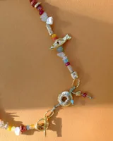 Collar corto de estilo artesanal, compuesto por una variedad de piedras naturales en tonos tierra, verdes, rojos y amarillos, intercaladas con pequeñas perlas de río. Presenta dijes de bronce macizo con formas orgánicas, incluyendo una figura de pez y un dije circular central, unidos por un cierre de barra y argolla.