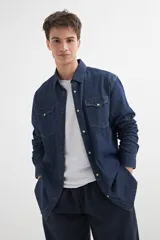 Camisa de denim azul con cuello clásico, cierre frontal con botones a presión, dos bolsillos con solapa abotonada en el pecho y mangas largas con puños abotonados.