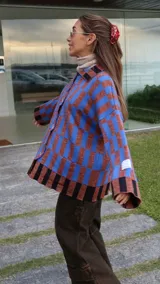 Buzo tipo saco abotonado con diseño de cuadros en tonos marrones y beige, corte recto y oversize. Presenta cuello camisero, cierre frontal con botones personalizados y dos bolsillos delanteros.