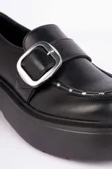 Mocasín negro de plataforma con suela chunky, tira frontal con hebilla metálica plateada y detalles de tachas en el borde del empeine.