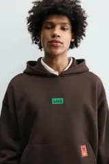 Sudadera con capucha de corte relajado, color marrón oscuro, con interior perchado. Presenta un bolsillo frontal tipo canguro, bordado con la palabra 'LOVE' en el pecho y un parche decorativo a contraste en el bolsillo. Posee puños y ruedo en rib.