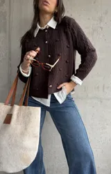 Cardigan de punto color chocolate, con diseño de trenzas y pompones decorativos. Presenta cuello redondo, cierre frontal con botones metálicos dorados y mangas largas.