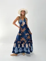 Vestido largo azul con estampado de palmeras y flores en tonos marrones y blancos. Tiene escote en V, espalda descubierta con elástico y tiras para atar al cuello.