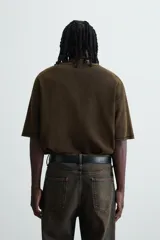 Camiseta de punto de algodón con corte boxy fit, cuello redondo y manga corta. Presenta un estampado frontal con efecto desgastado en tonos blanco y rojo sobre un fondo marrón oscuro/verde oliva lavado.
