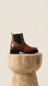 Bota tipo Chelsea de cuero color marrón, con paneles laterales elásticos en color negro y suela track robusta de goma negra.