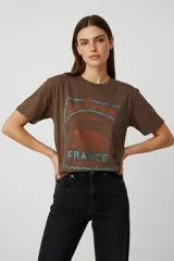 Remera de algodón color marrón con cuello redondo y mangas cortas. Presenta un estampado frontal con diseño gráfico estilo vintage que incluye texto y una ilustración de paisaje.
