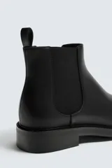 Bota corta estilo Chelsea de cuero liso negro, con elásticos laterales y tirador trasero para calce. Presenta horma redonda y suela con ligero tacón.