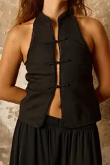 Chaleco halter negro confeccionado en lino con lurex, de escote alto y abierto en el frente con cierre de alamares y terminaciones con vivos de mostacillas.