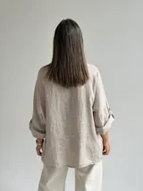 Blusa holgada de lino color beige, con escote en V, mangas tres cuartos con trabilla y bolsillos delanteros aplicados con bordes deshilachados. Cuenta con tiras frontales decorativas con botones de madera.