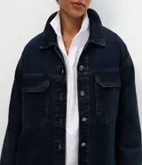 Campera tipo overshirt de denim en color azul oscuro, con corte amplio y largo extendido. Presenta cuello clásico, cierre frontal con botones metálicos, mangas largas y dos bolsillos delanteros con solapa.