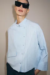 Camisa de corte oversize con estampado de rayas verticales, cuello redondo tipo mao y cierre frontal con botones. Presenta manga larga con puños anchos, un bolsillo de plastrón con solapa en el frente y bajo asimétrico.