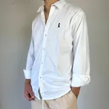 Camisa de manga larga color beige claro, con cuello clásico, cierre frontal con botones y logo bordado en el pecho. Corte regular.