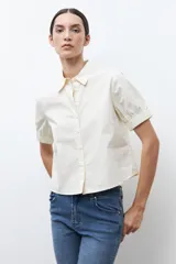 Camisa de algodón color beige claro, con cuello camisero, botones frontales y mangas cortas abullonadas con elástico en el puño.