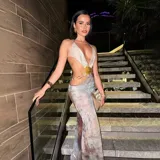 Vestido largo de fiesta con escote halter, espalda descubierta y abertura lateral en la falda. Presenta un top triangular con detalle de abertura en el abdomen y una cadena dorada con dije de concha que rodea la cintura. El estampado es abstracto en tonos grises y marrones.