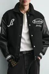 Cazadora estilo varsity con cuerpo de tejido con lana y mangas de efecto piel. Presenta cuello solapa, cierre frontal con botones a presión, bolsillos laterales y puños con elástico a rayas. Incluye parches y bordados decorativos en el frente.