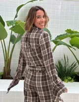 Camisa de corte holgado con estampado de cuadros en tonos marrones y beige. Presenta cuello clásico, cierre frontal con botones y mangas largas con aberturas laterales que permiten anudar la prenda en la cintura.