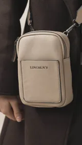 Bandolera pequeña de diseño compacto en color mostaza, confeccionada en material sintético con textura granulada. Presenta un bolsillo frontal con el logo de la marca impreso, cierre superior y correa ancha ajustable y desmontable.