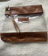 Necessaire de diseño compacto confeccionado en cuero color marrón combinado con material sintético transparente. Presenta cierre superior, costuras reforzadas y una etiqueta frontal de cuero con el logo de la marca grabado.